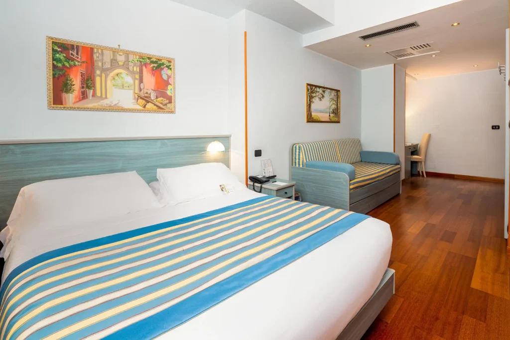 immagine di una camera classic, con pavimento in legno, letto matrimoniale in primo piano, divano sullo sfondo e colori che richiamano l'azzurro del mare