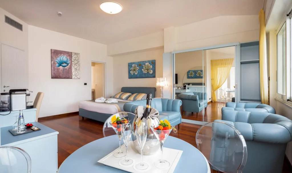 nell'immagine, l'interno di una suite con pavimento in legno, 3 poltrone in pelle azzurra, in primo piano un tavolino con delle coppe di frutta fresca e una bottiglia di champagne e, sullo sfondo, un letto matrimoniale