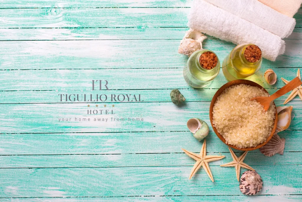 nell'immagine si vedono elementi di una Spa: una ciotola con dei sali per lo scrub, due boccette di oli essenziali, degli asciugamani arrotolati e delle conchiglie