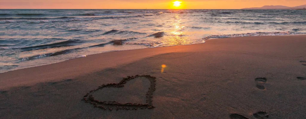 nell'immagine un bagnasciuga al tramonto, con un cuore disegnato nella sabbia